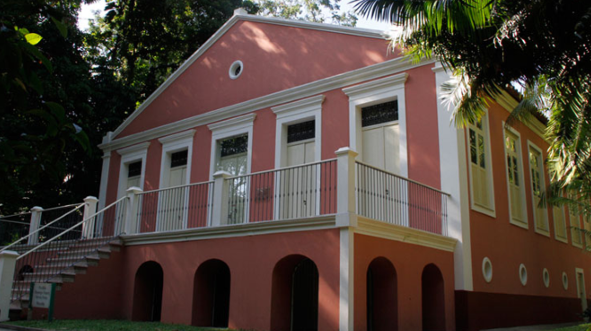 Prédio histórico do Museu Paraense Emílio Goeldi em tons de rosa, com varanda, escadaria e arcos, cercado por vegetação.